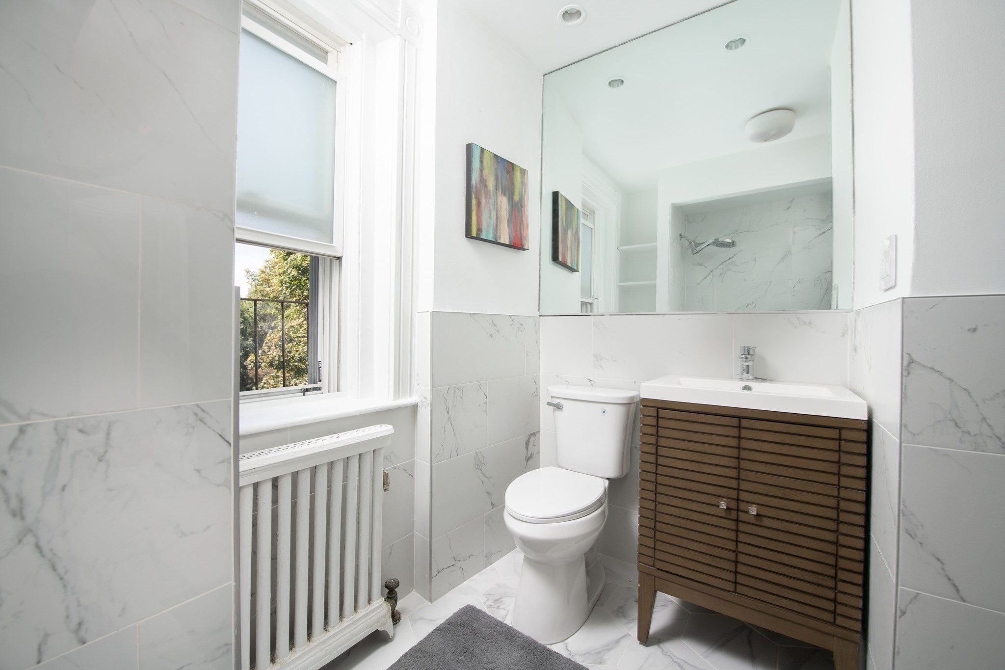 Renovated bathroom with marble-look tile, wood slatted vanity, and a window above a radiator.