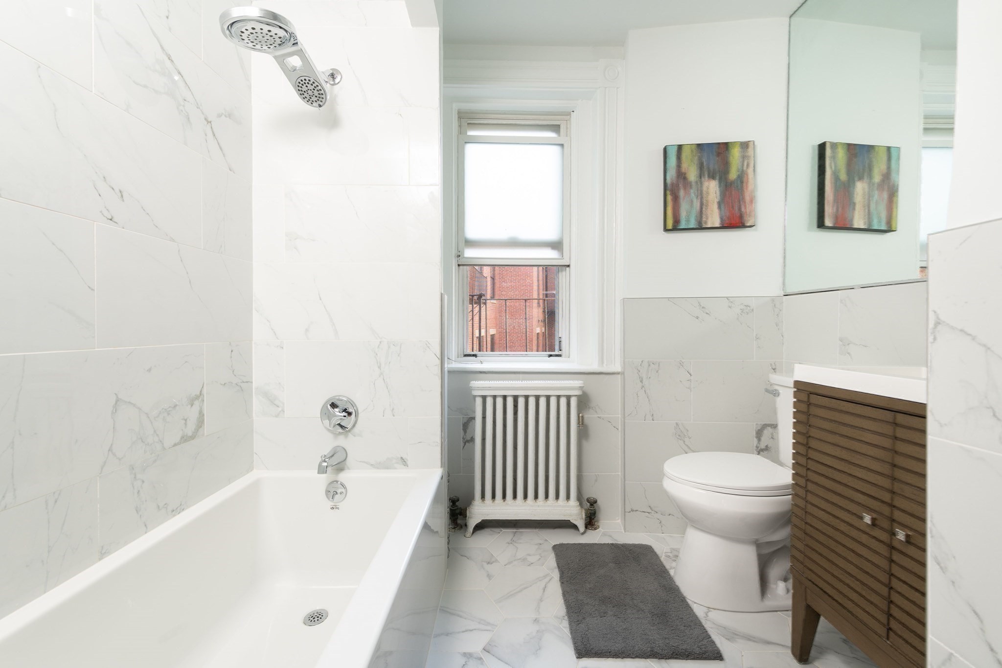 Bathroom with marble-look tile, bathtub and shower, radiator beneath a window, and wood-front vanity.