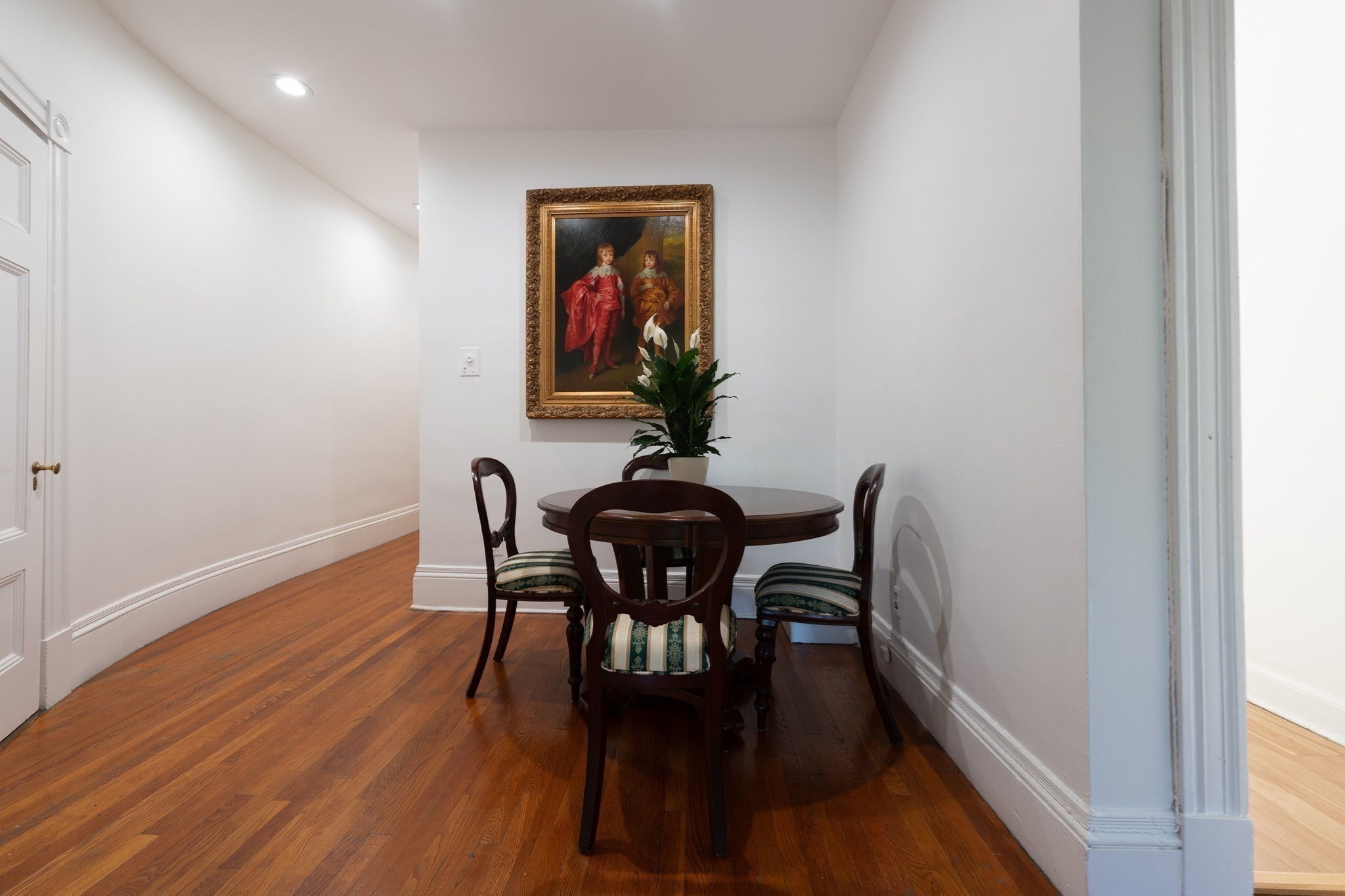 Dining nook with a round dark wood table, hardwood floors, and framed artwork above a potted plant.