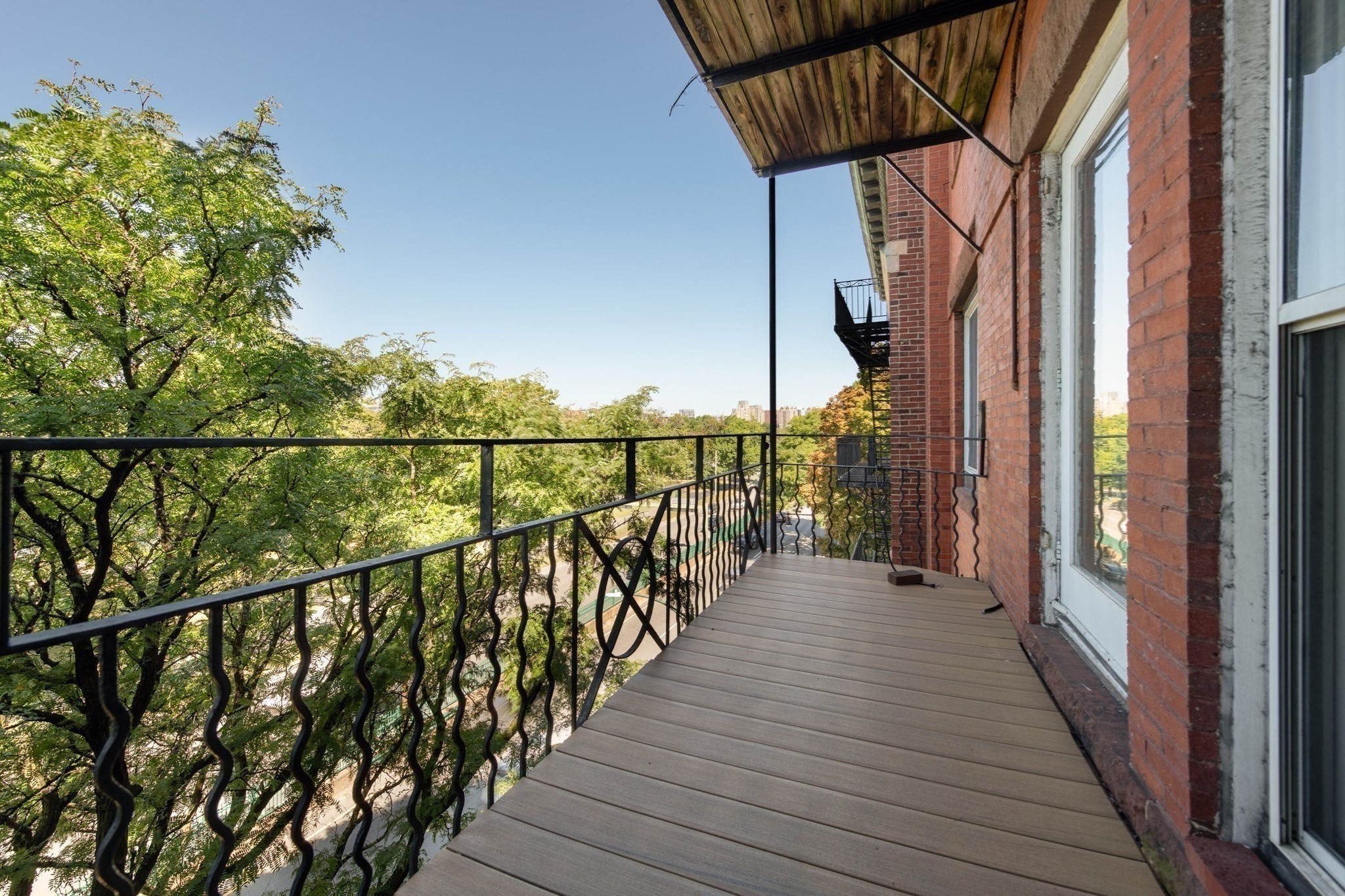 Balcony with a wood-plank awning and black railing overlooking leafy treetops and distant buildings.