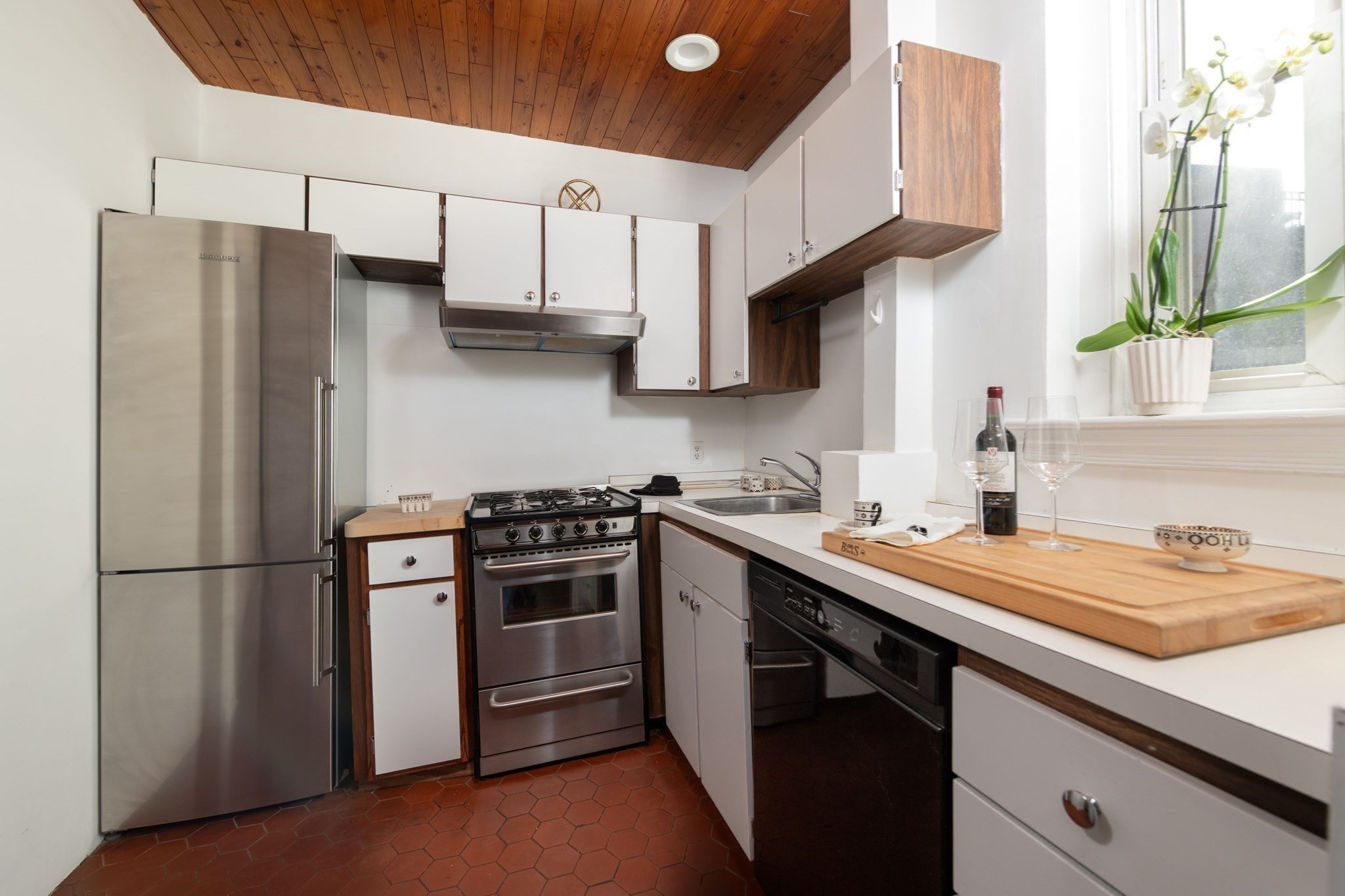 Compact kitchen with stainless steel appliances, terracotta tile floor, wood ceiling, and a bright window.