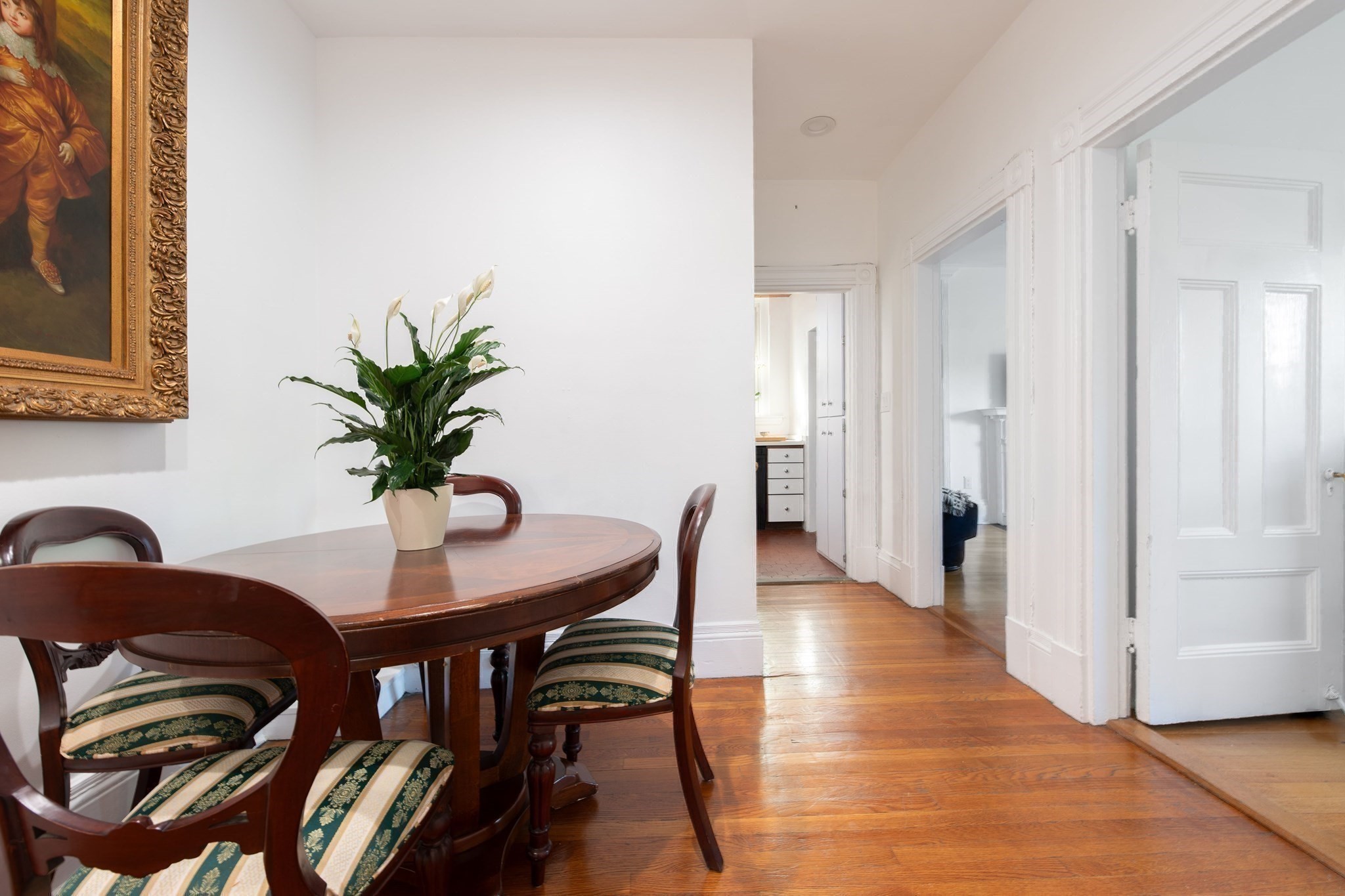 Dining area with a round wooden table, upholstered chairs, and a potted plant beside a hardwood hallway.