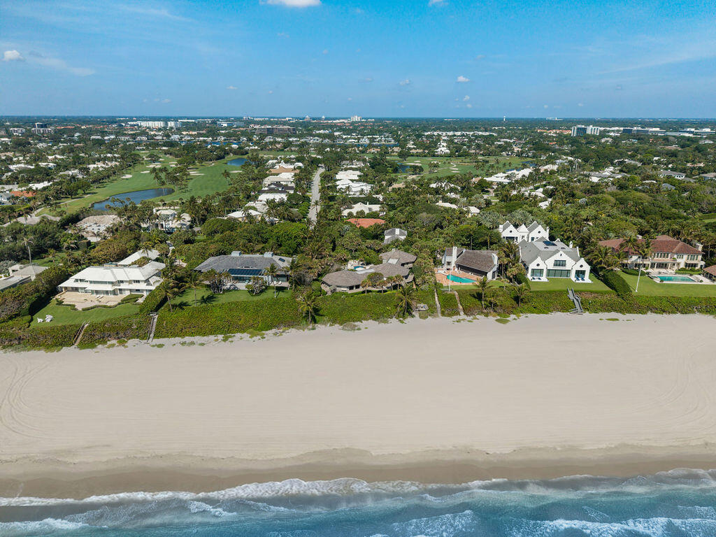 Aerial view of beachfront homes, sandy shoreline, ocean waves, and green golf course grounds inland.