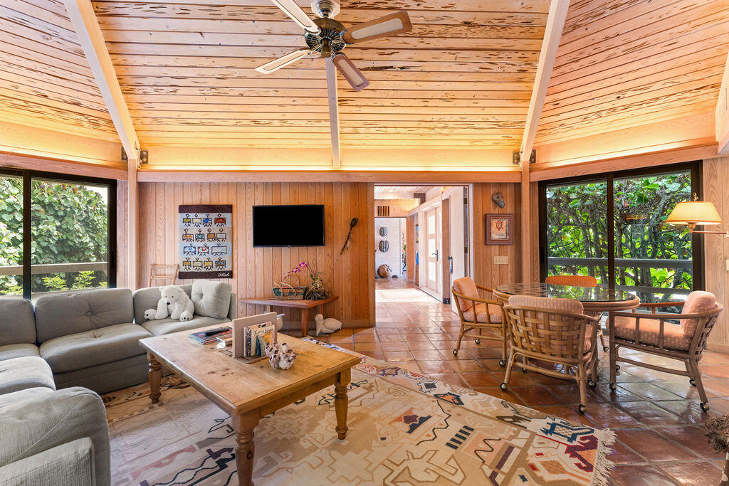 Living room with a vaulted wood ceiling, gray sectional, patterned rug, and dining area beside large windows.