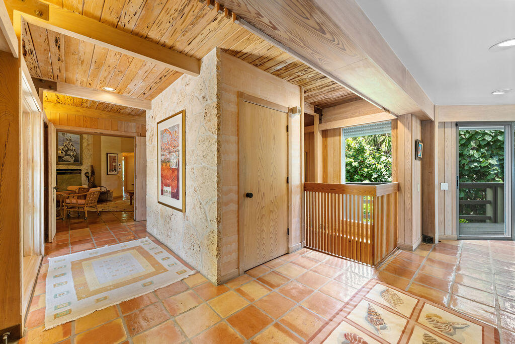 Entry hall with terracotta tile floors, a wood-beamed ceiling, stone wall, and railing beside leafy windows.