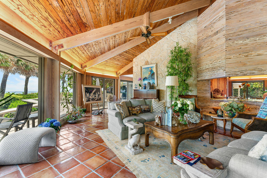 Living room with vaulted wood ceiling, terracotta floors, stone wall, and ocean views through patio windows.