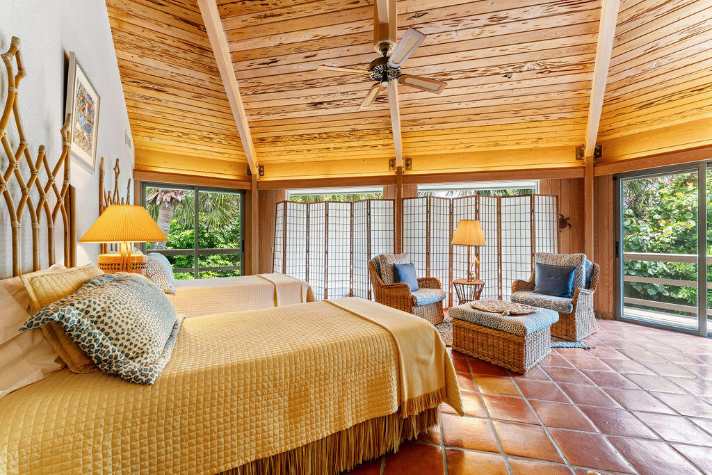 Bedroom with two beds, wicker seating, a wood-plank ceiling, and windows overlooking greenery.