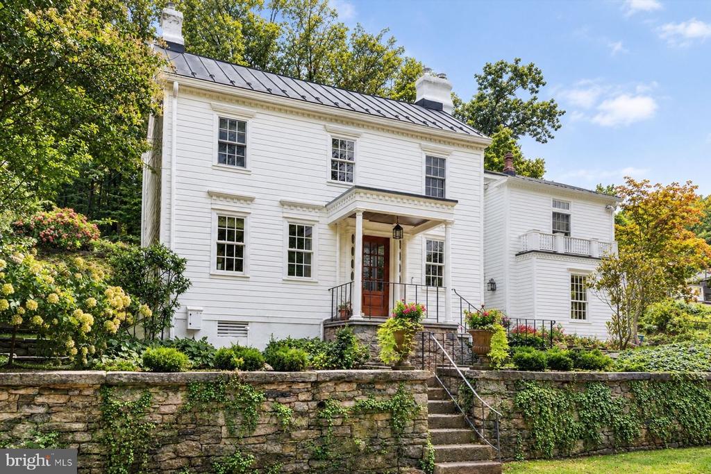 White two-story house with a dark metal roof, covered entrance, and stone garden wall.