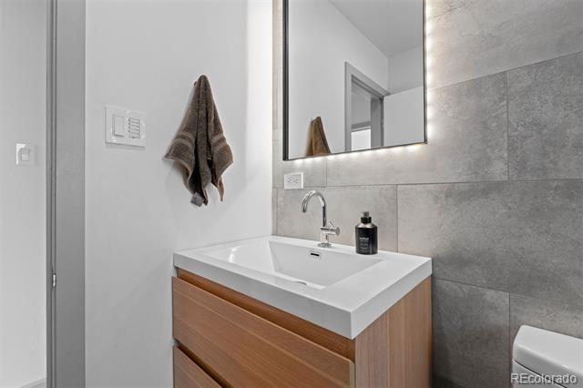 Bathroom vanity with a white rectangular sink, wood-front cabinet, and gray tiled wall.