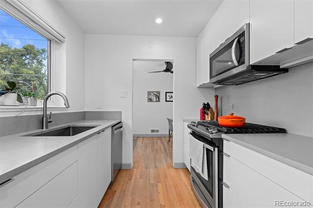 Modern white kitchen with wood floors, stainless-steel appliances, and a window overlooking trees.
