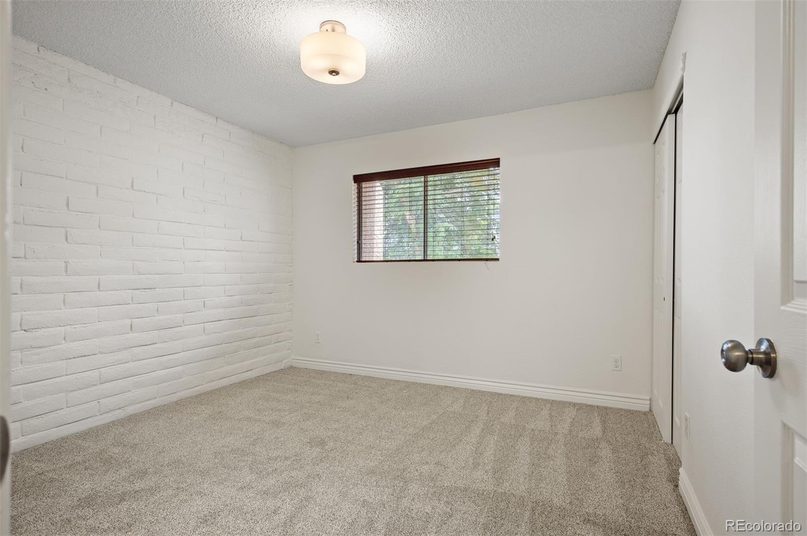 Empty bedroom with beige carpet, a white brick accent wall, and a window with blinds.