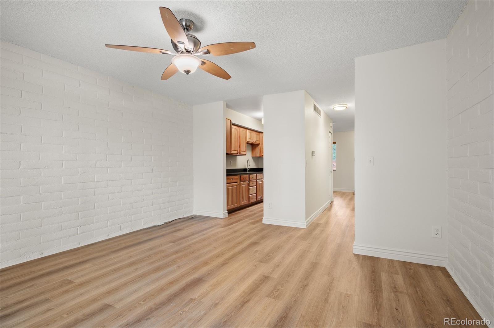 Empty living room with white brick walls, wood-look floors, ceiling fan, and kitchen beyond.