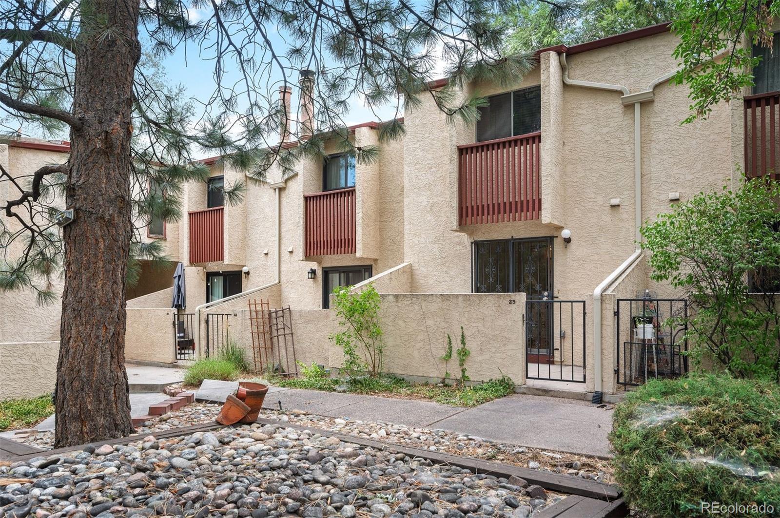 Beige stucco condominium exterior with a gated patio, red-brown balcony railings, and mature trees.