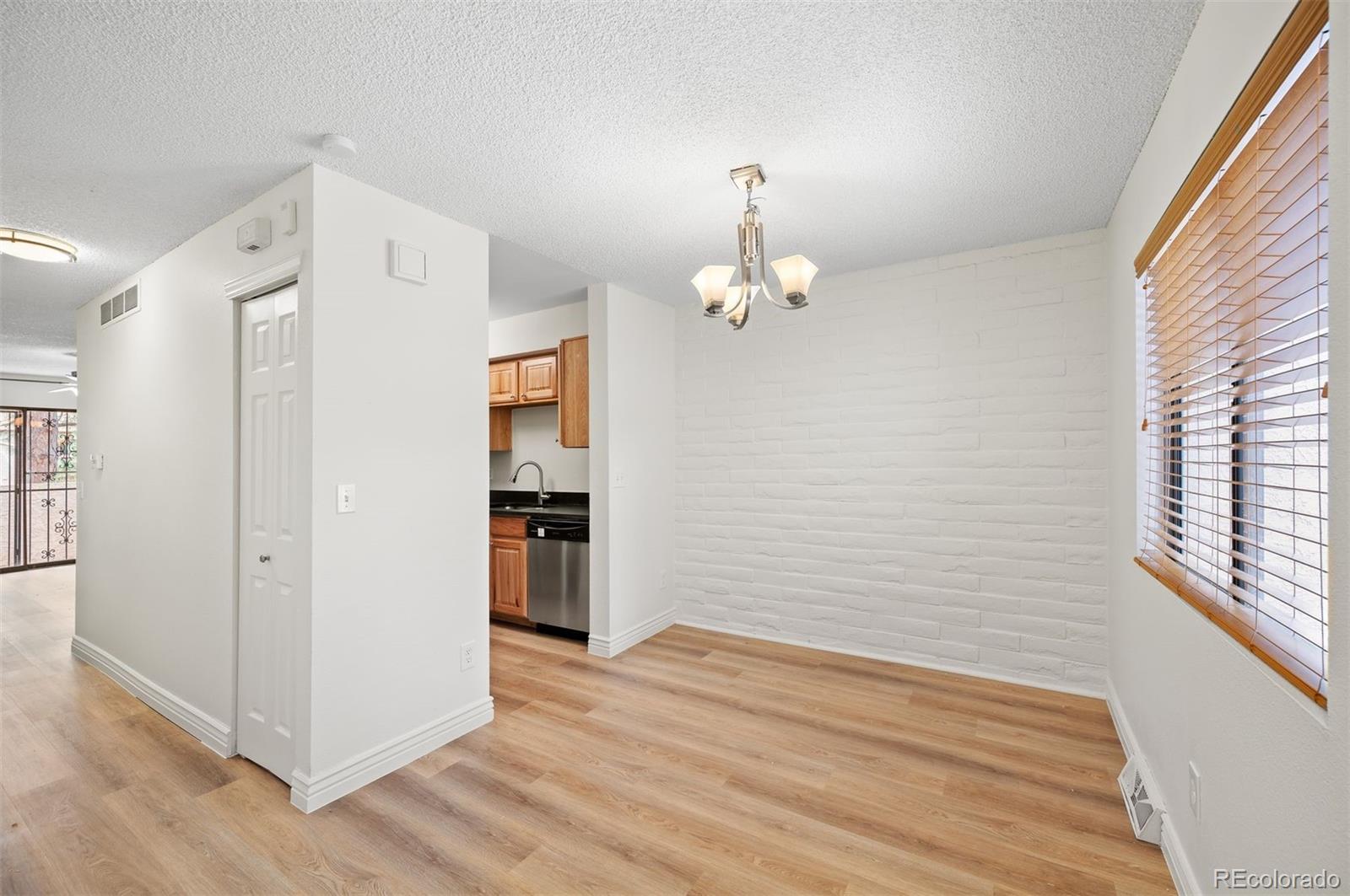 Vacant dining area with white brick accent wall, wood flooring, chandelier, and kitchen opening.