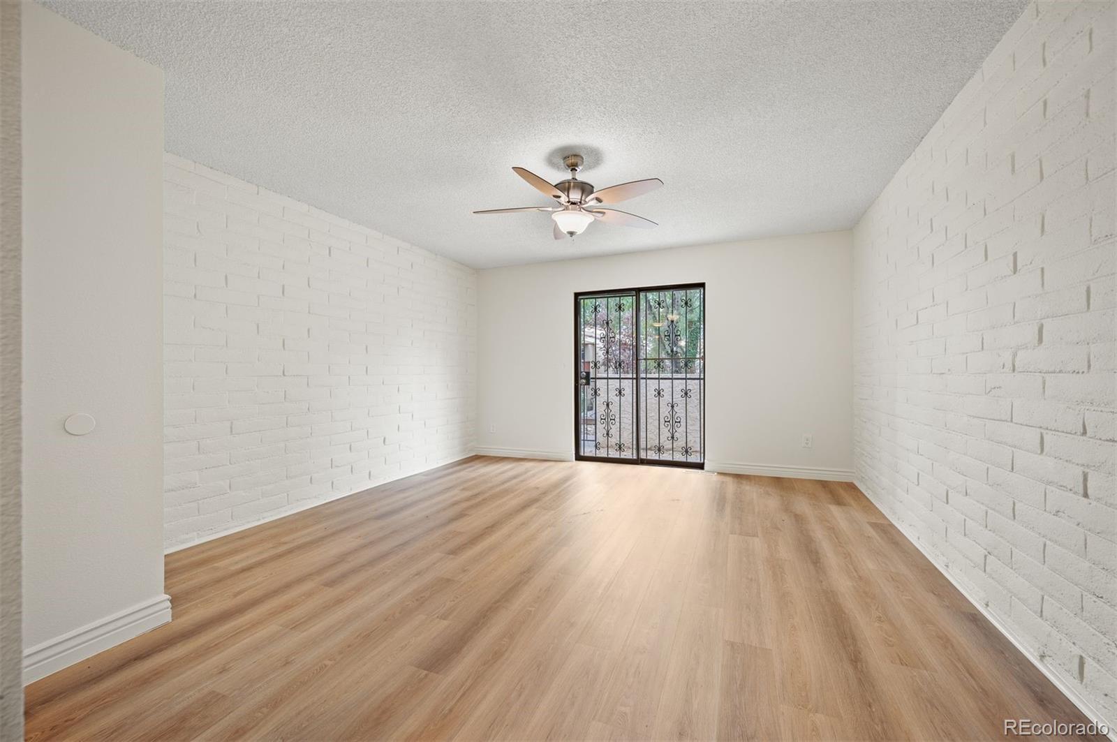 Empty living room with white brick walls, wood floors, ceiling fan, and barred glass patio door.