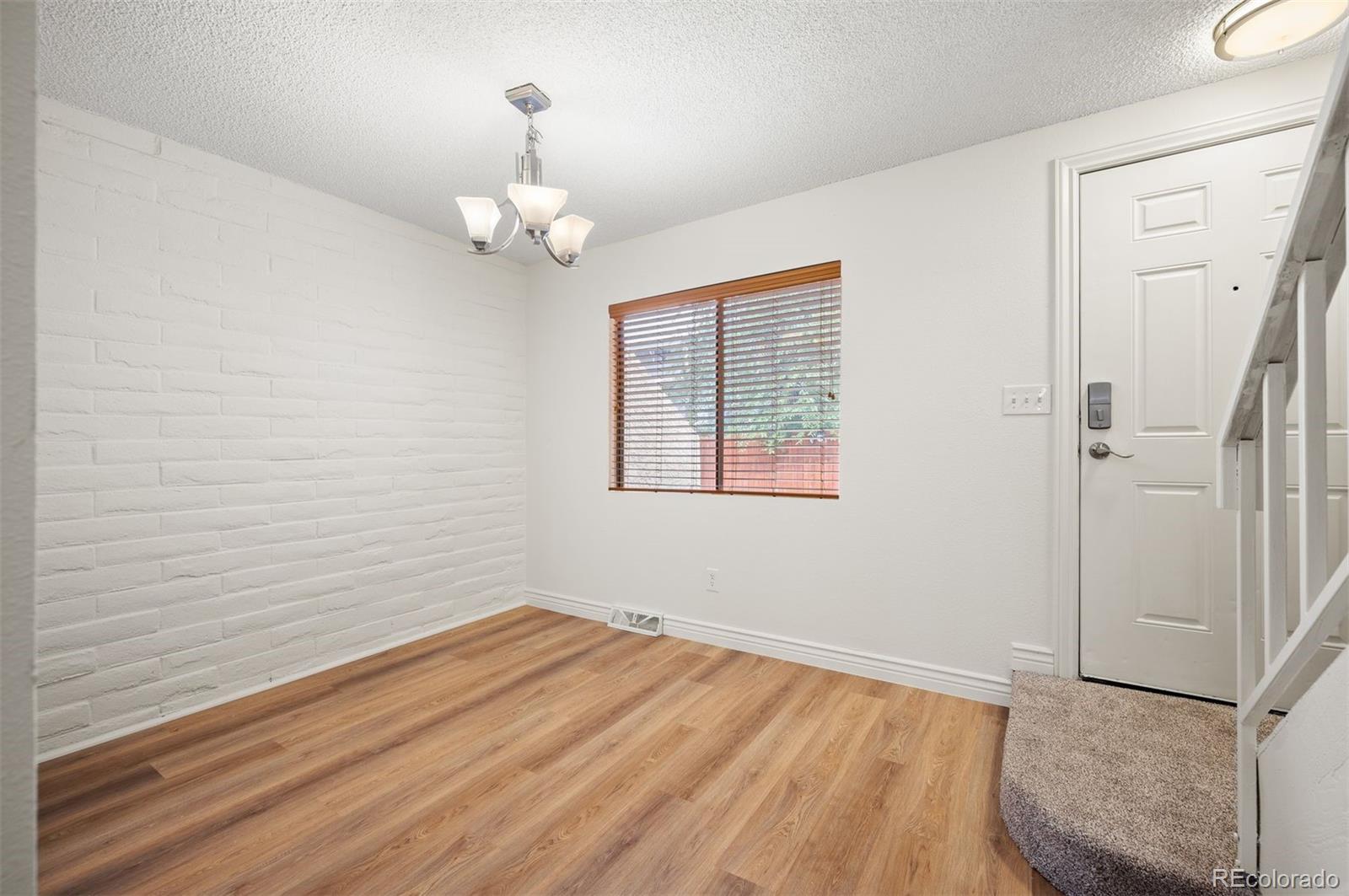 Empty room with wood-look flooring, a white brick wall, window blinds, and an entry door beside carpeted stairs.