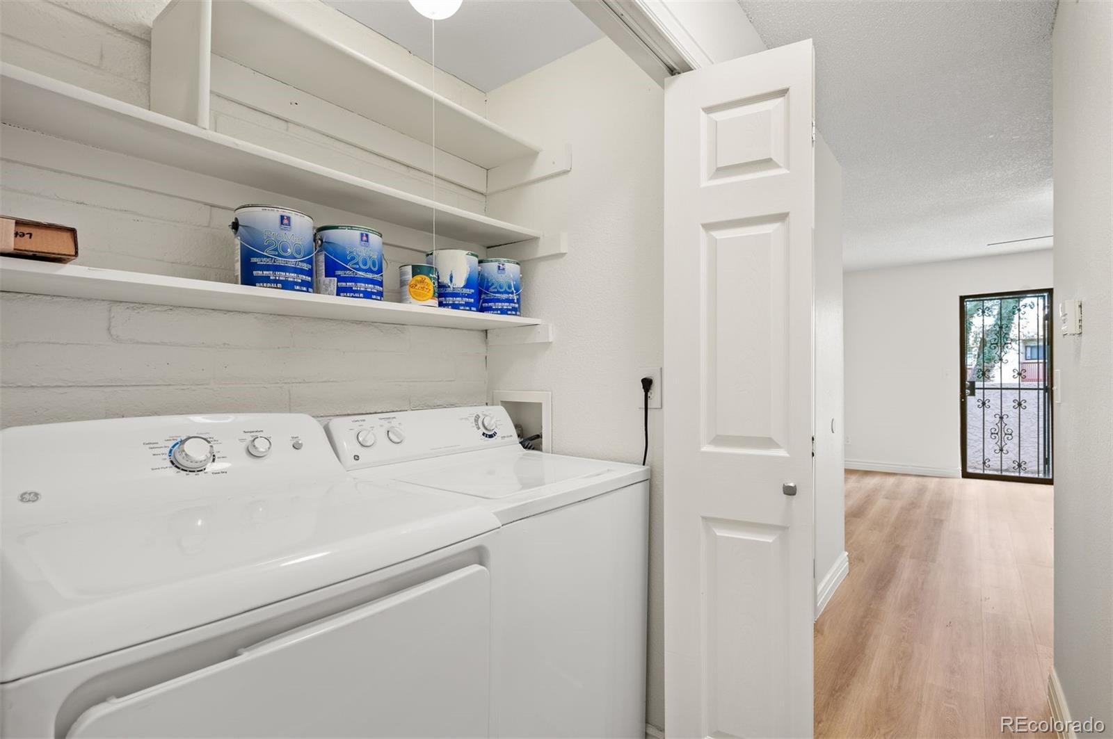Laundry area with white washer and dryer, open shelves, and doorway to a wood-floored hall.