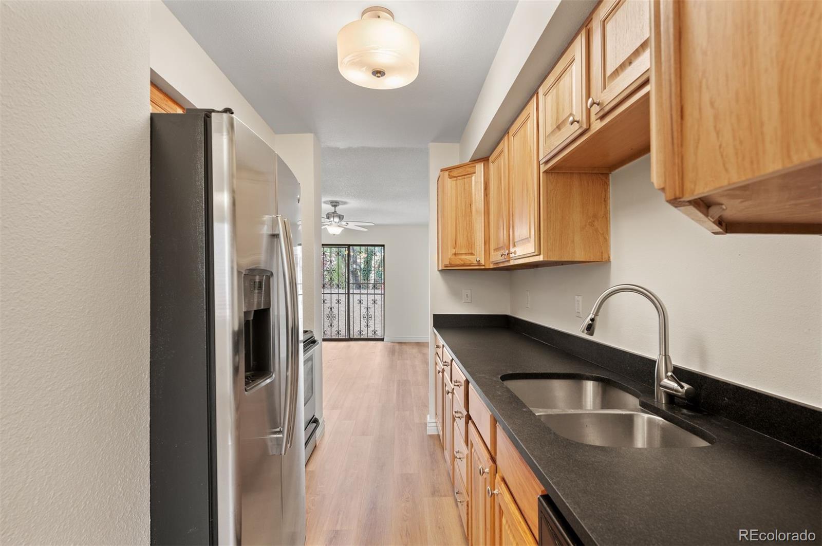 Galley kitchen with wood cabinets, black countertops, stainless steel refrigerator, and double-basin sink.