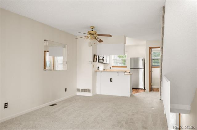 Unfurnished living room opening to a kitchen, with beige carpet and a ceiling fan.