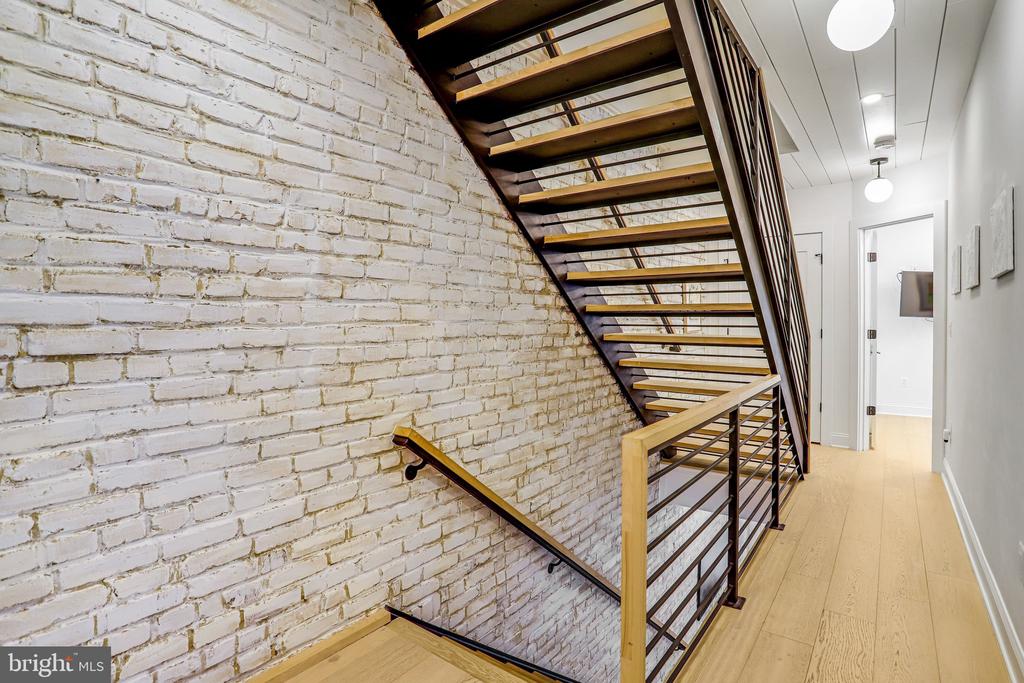 Interior staircase beside a white-painted brick wall, with wood treads and dark metal railings.