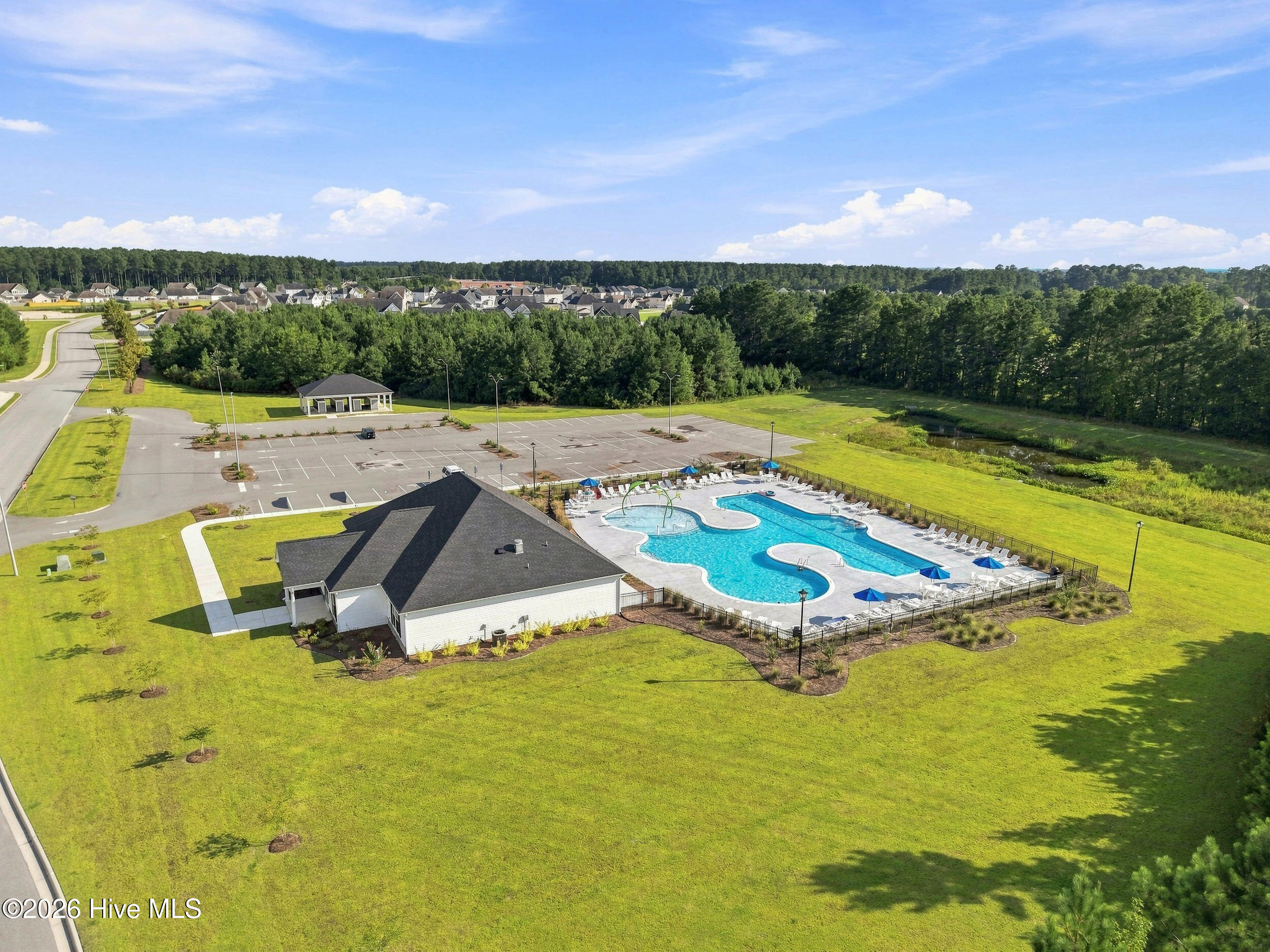 Aerial view of a community pool and clubhouse surrounded by lawns, parking, and trees.