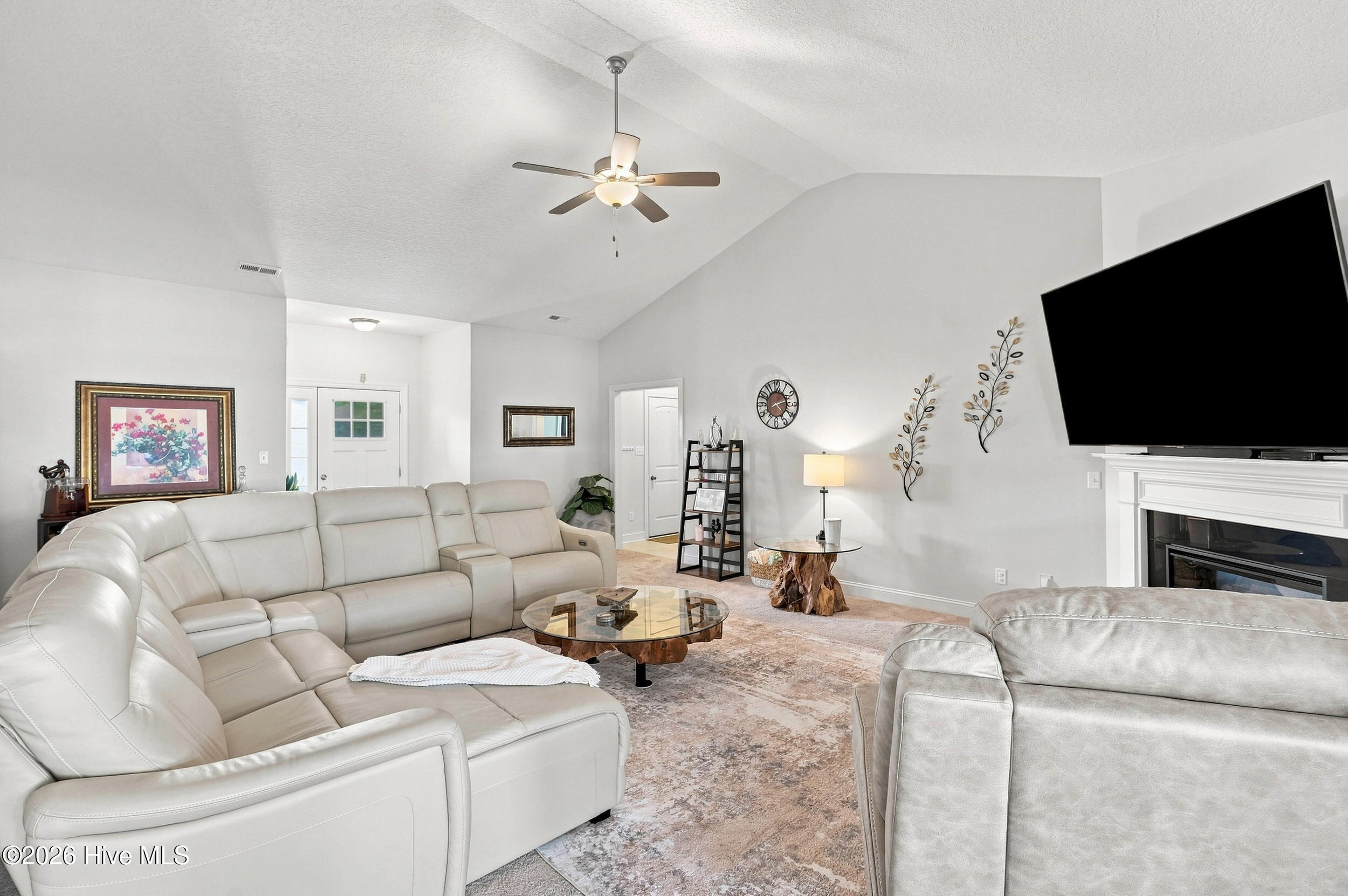 Living room with white sectional sofas, vaulted ceiling, and a wall-mounted television above the fireplace.