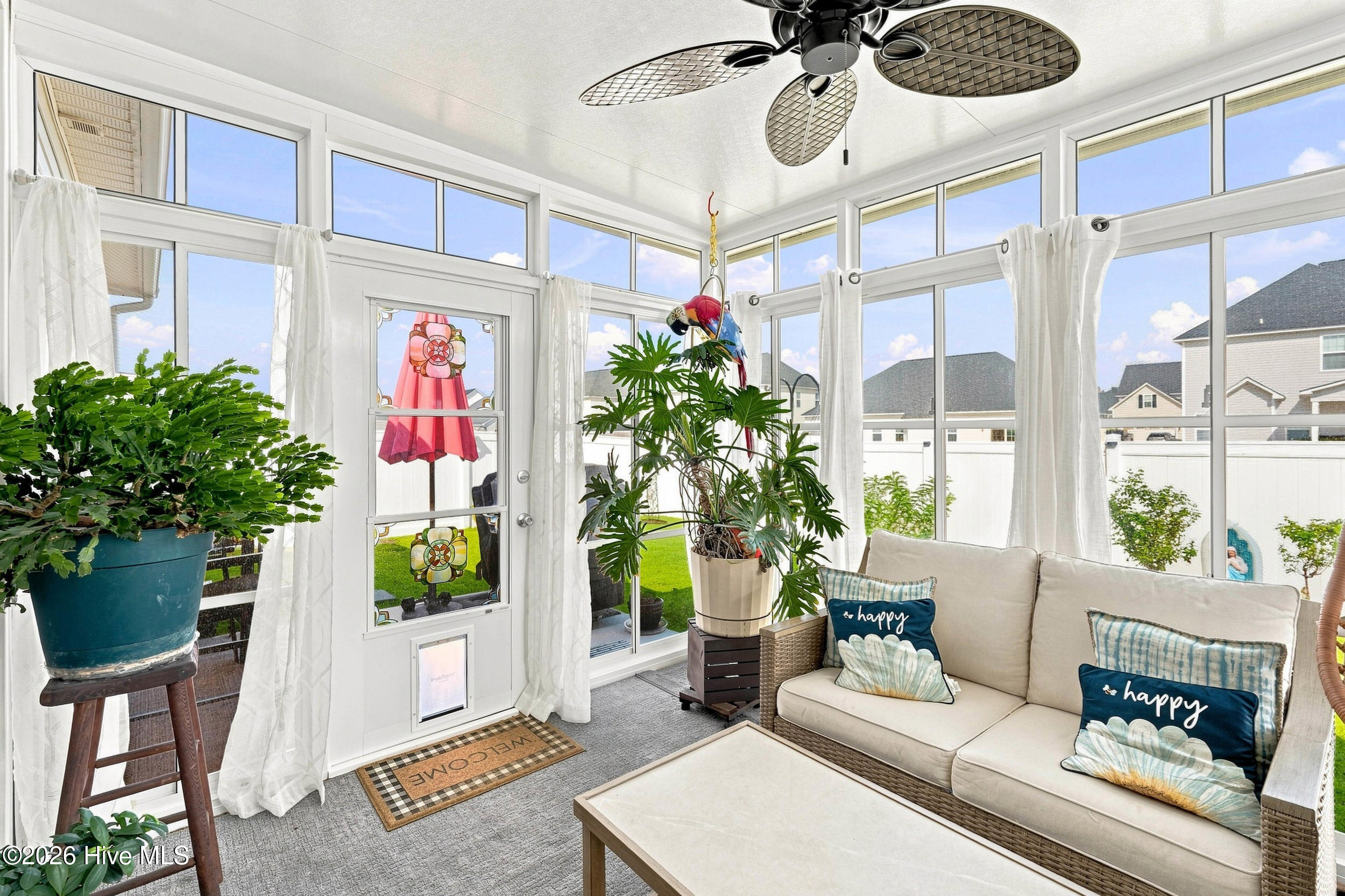 Furnished sunroom with tall windows, a wicker sofa, leafy plants, and a ceiling fan.