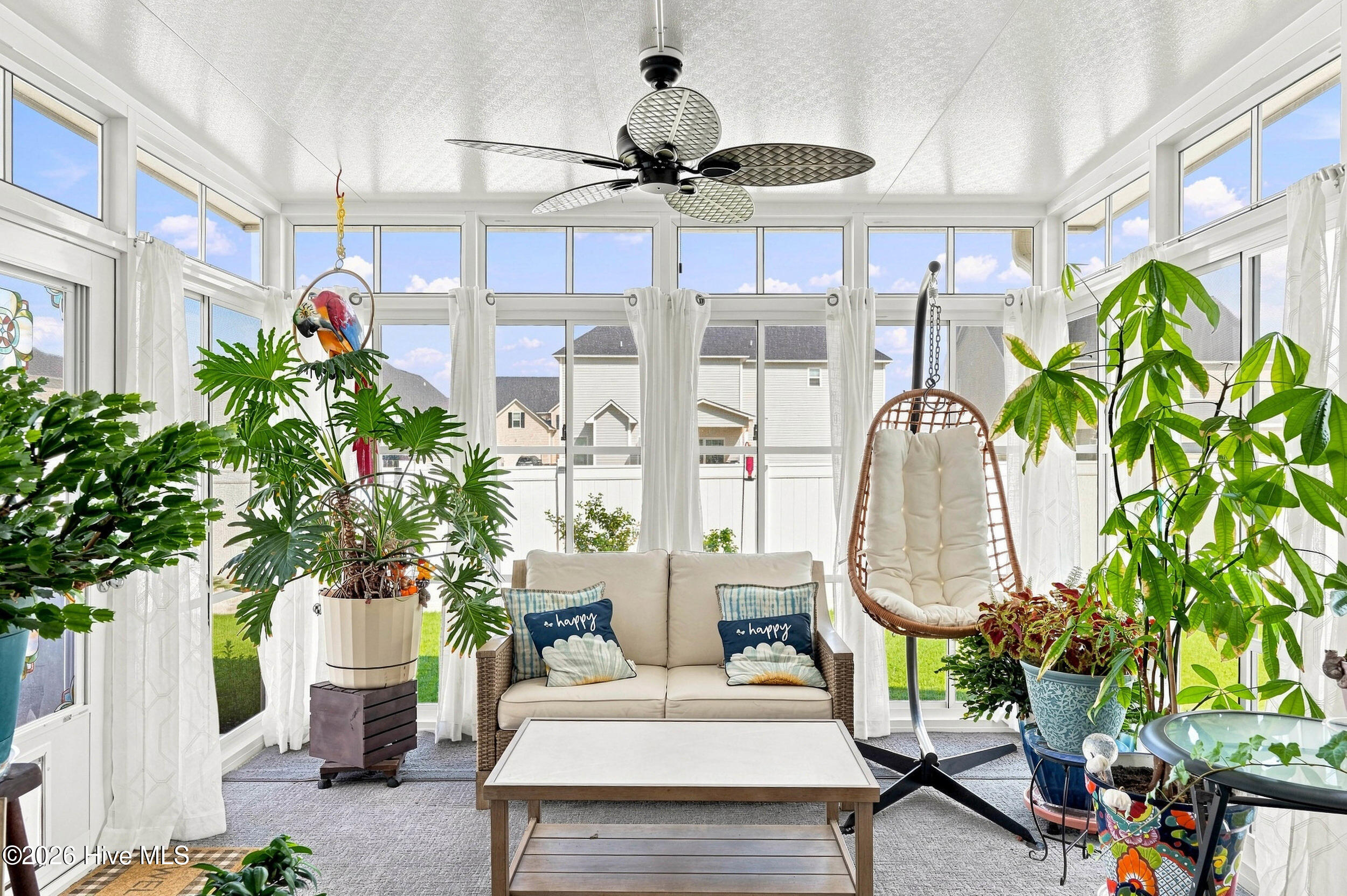 Bright sunroom with floor-to-ceiling windows, a cushioned sofa, hanging chair, and abundant plants.