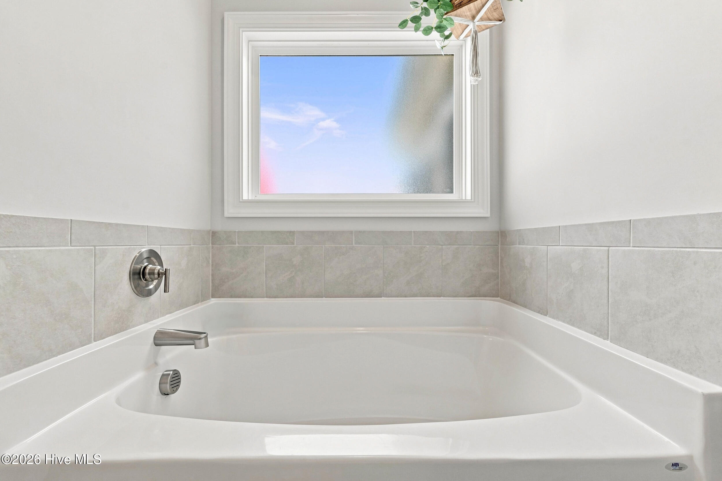 White bathtub with chrome fixtures beneath a window, surrounded by light gray tile.