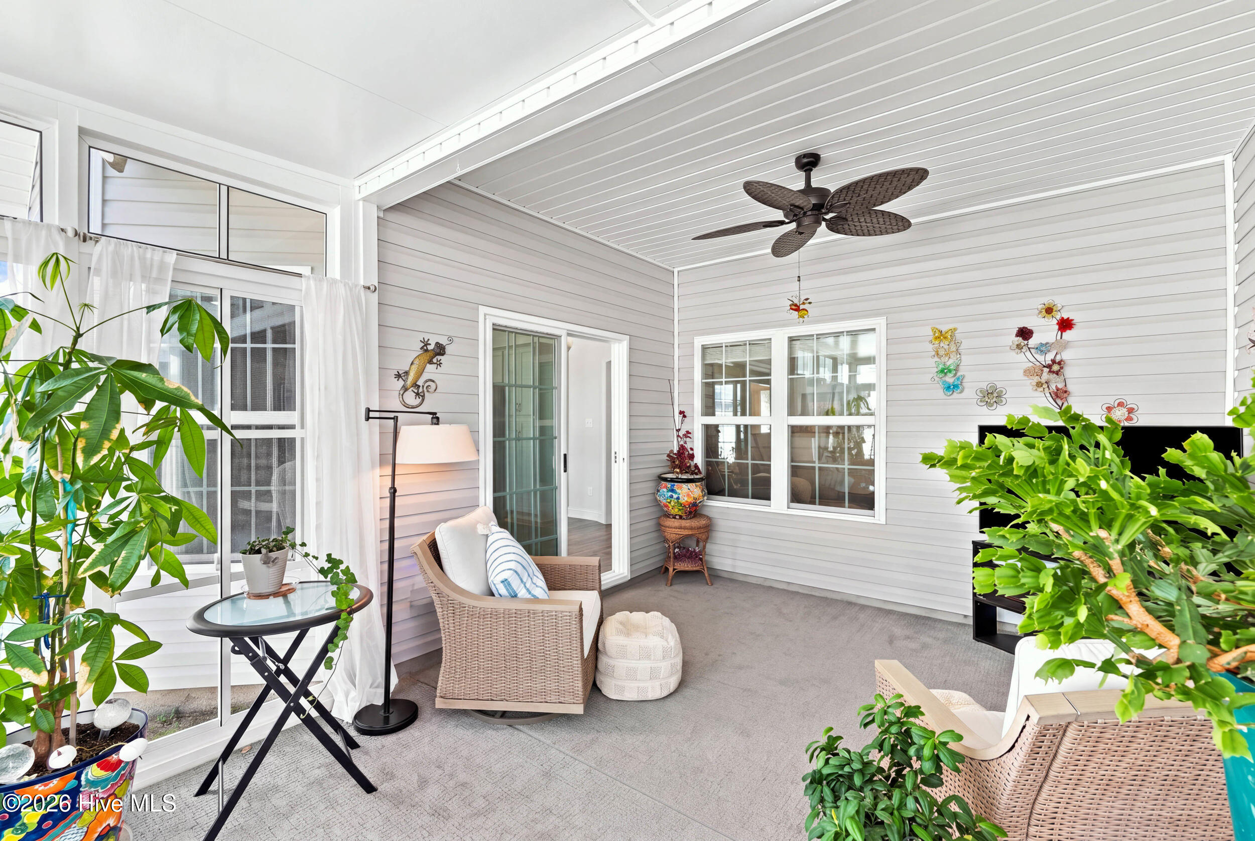 Covered patio with wicker seating, a ceiling fan, large windows, and leafy plants.