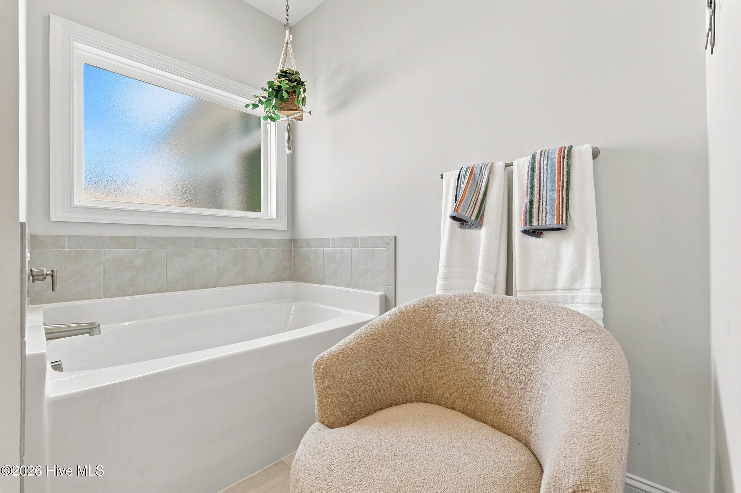 Bathroom with a white bathtub, frosted window, striped towels, and a beige upholstered chair.