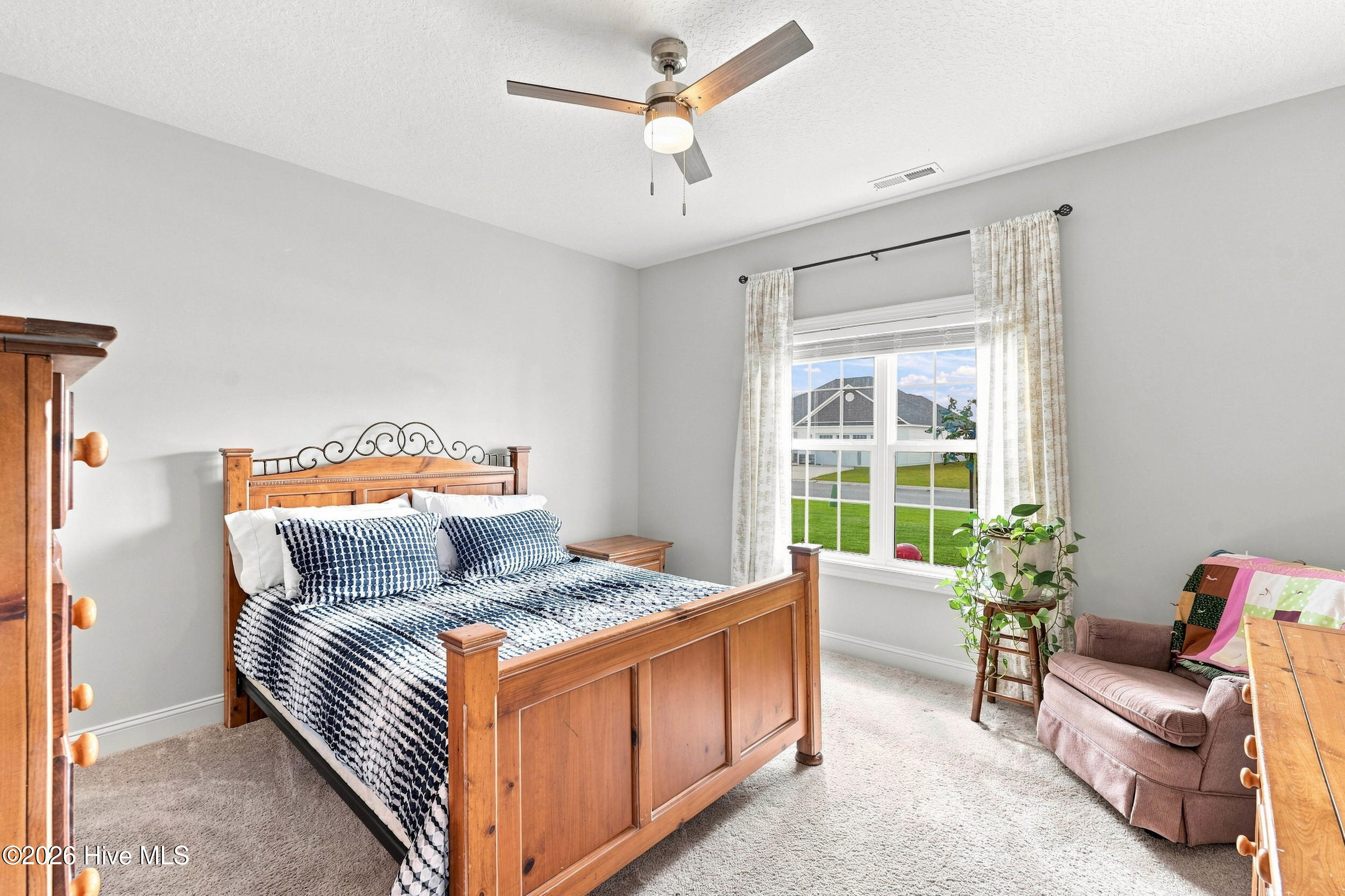 Bedroom with a wooden bed, patterned bedding, ceiling fan, and a curtained window overlooking a lawn.
