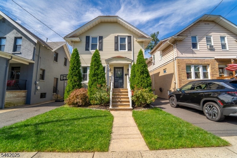 Two-story beige house with a covered entry, evergreen shrubs, and a walkway through the front lawn.