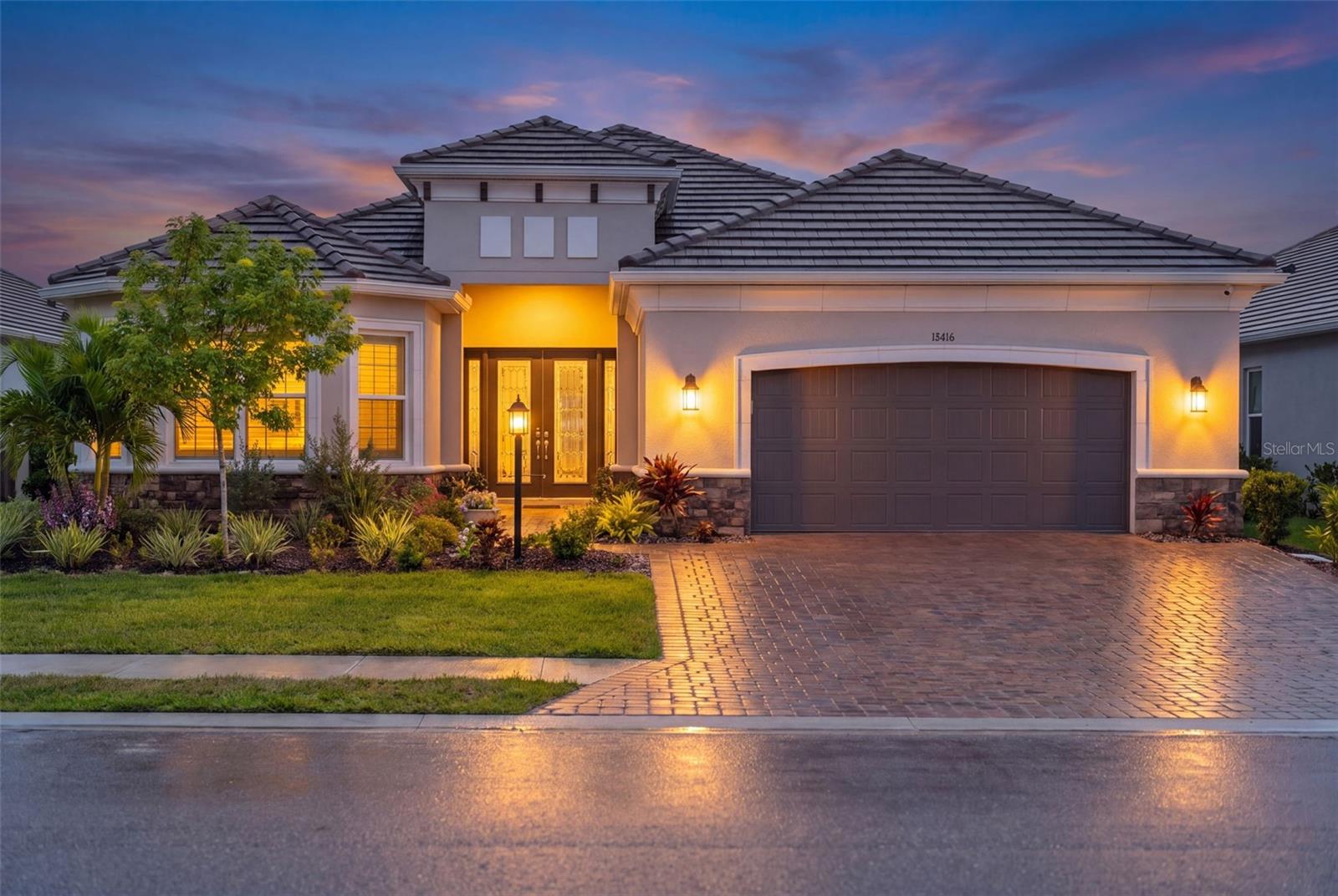 Stucco home at dusk with an illuminated entry, landscaped yard, and wet paver driveway.