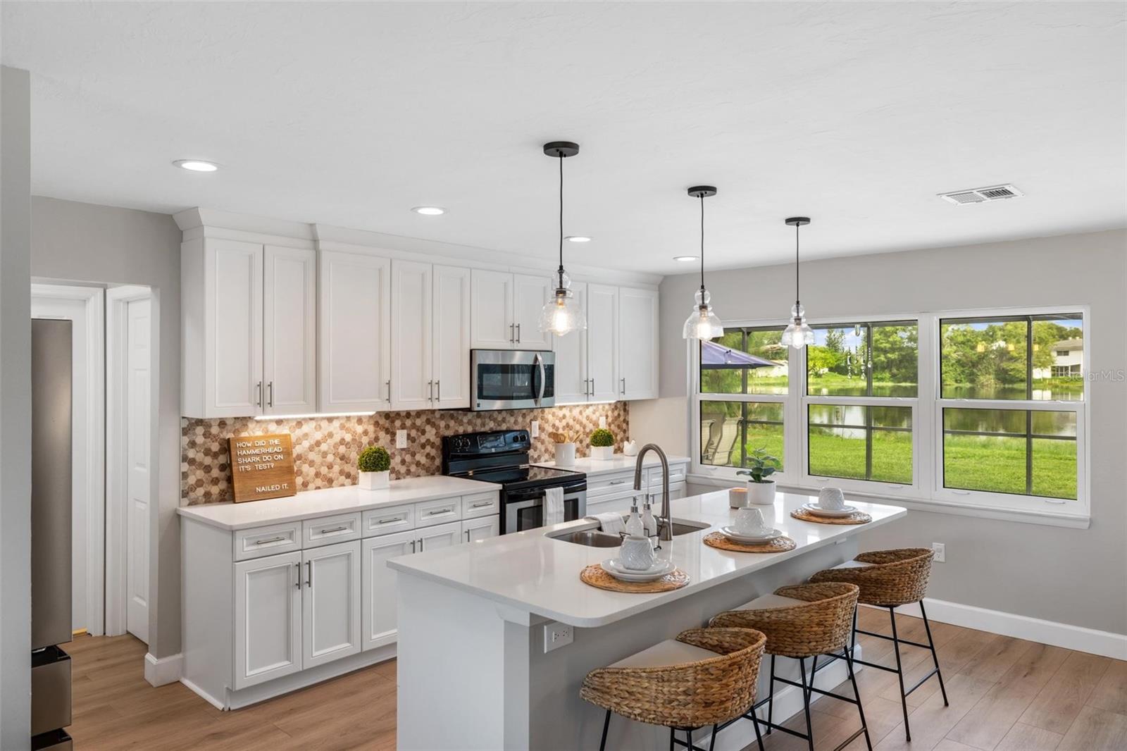 Bright white kitchen with a long island, woven bar stools, and windows overlooking a pond.