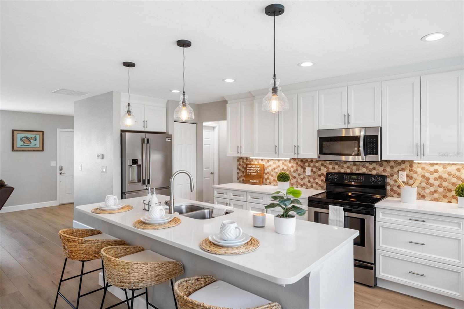 Bright kitchen with a large white island, woven stools, stainless refrigerator, and patterned backsplash.