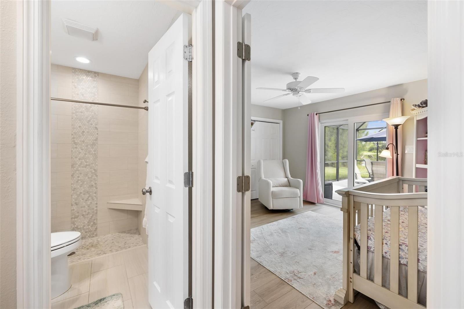Bedroom with a white crib and armchair beside sliding glass doors, viewed through an open tiled bathroom.