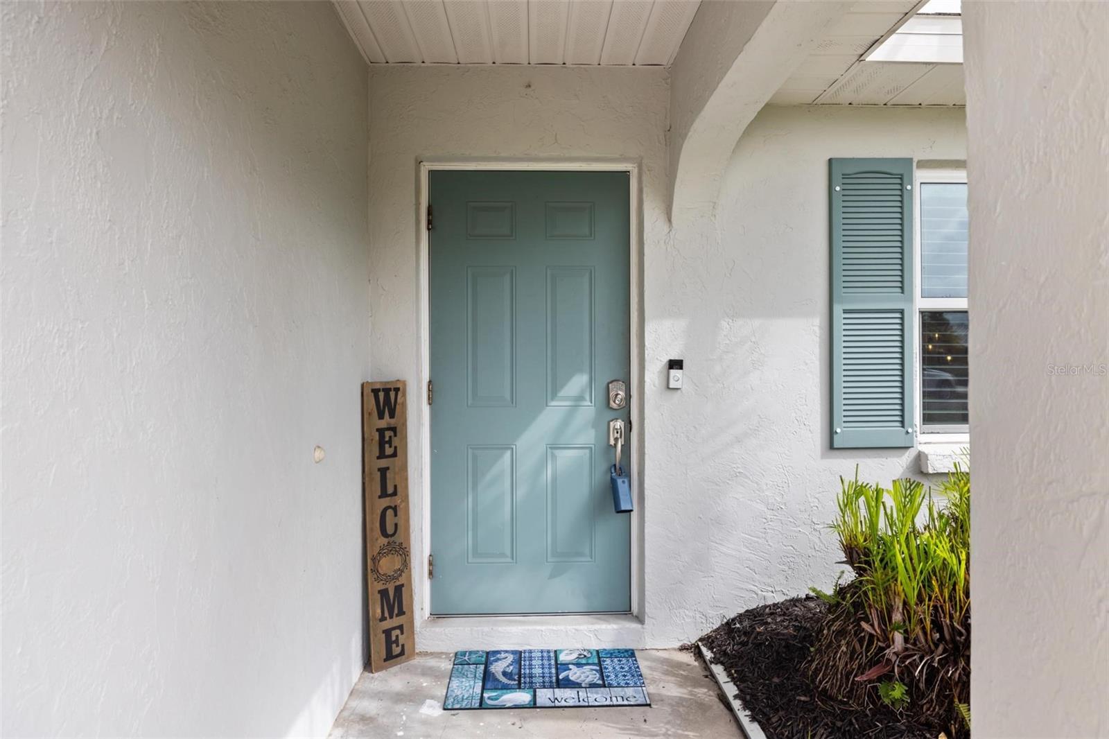 Covered home entry with a teal front door, matching shutters, and tropical landscaping.