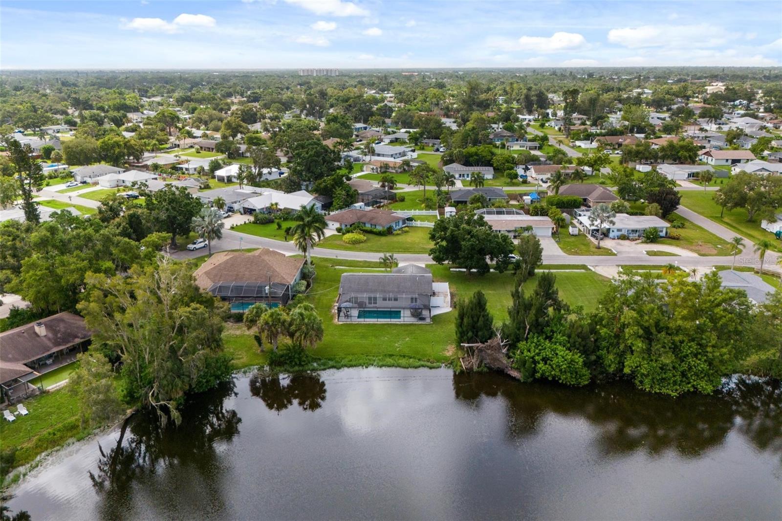 Aerial view of a Venice home beside a pond, with a screened pool and tree-lined neighborhood.