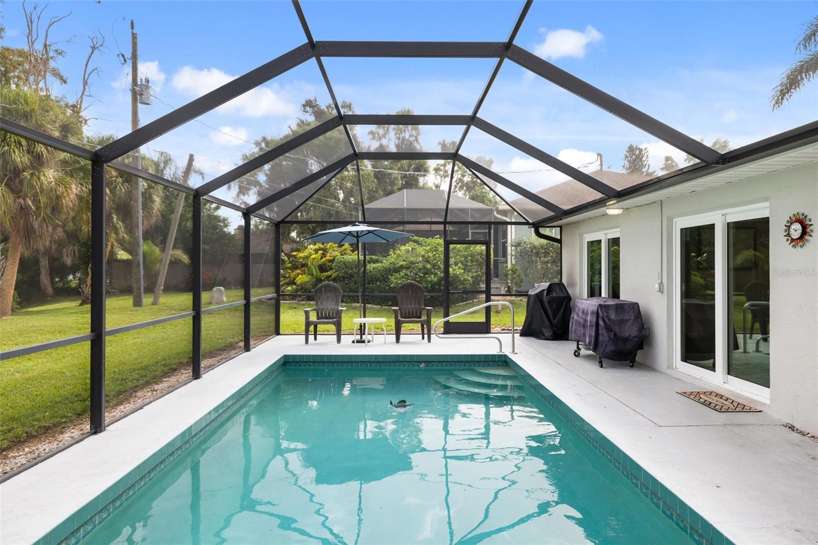Backyard pool beneath a screened enclosure, with patio chairs and tropical landscaping beyond.