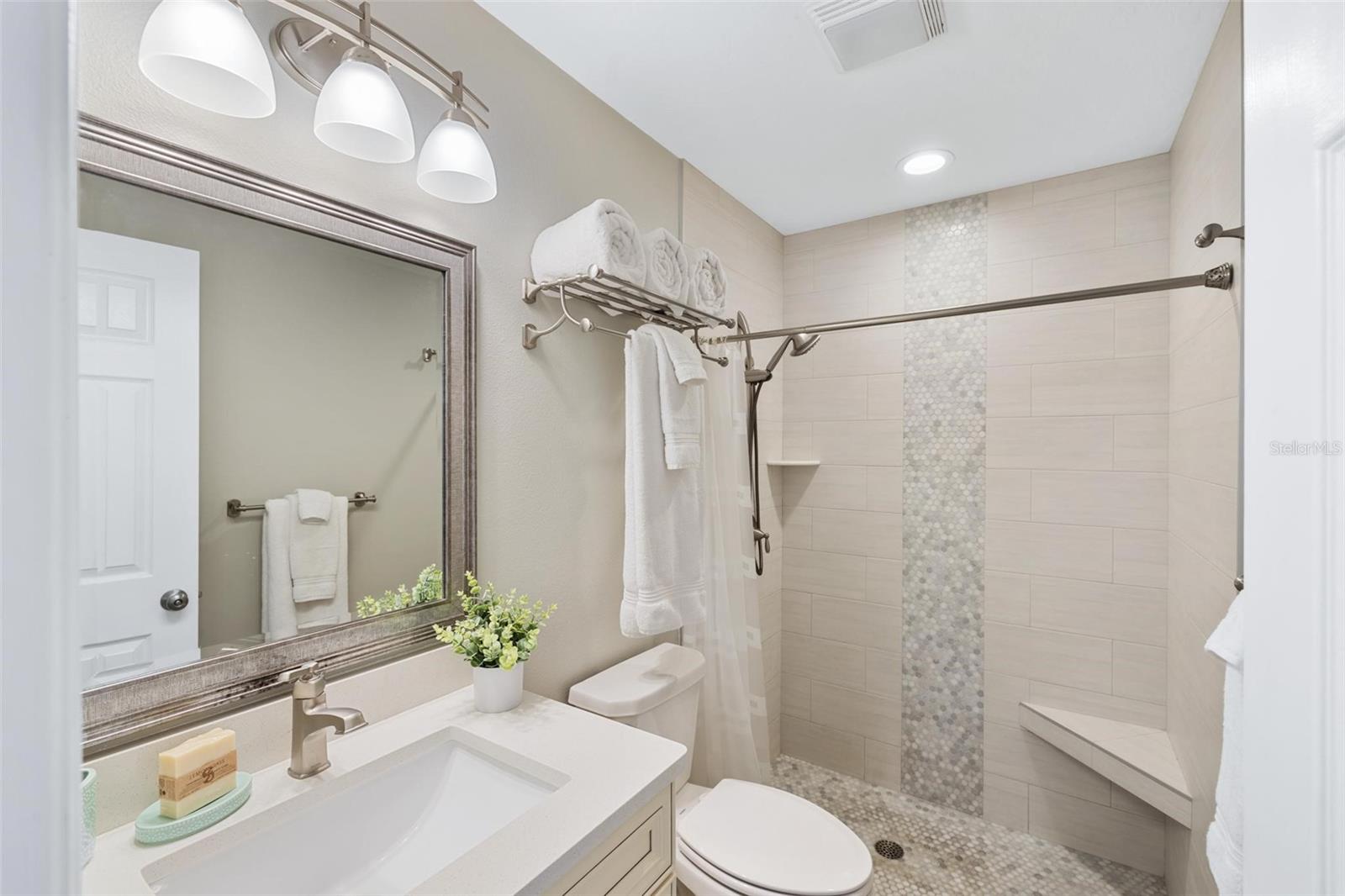 Bathroom with a white vanity and tiled walk-in shower featuring a pebble-mosaic accent strip and bench.