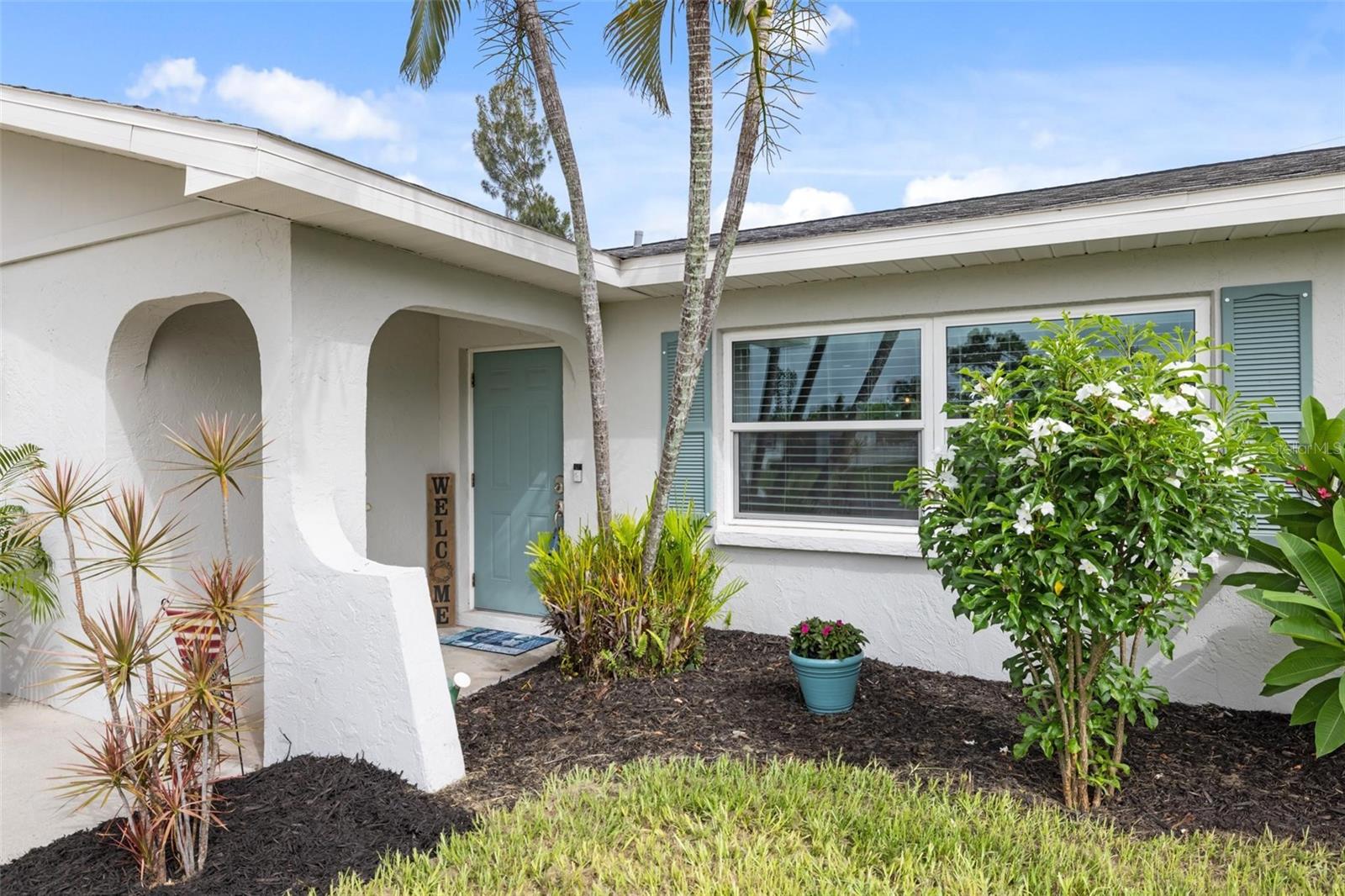 White stucco home entrance with a teal door, arched porch, and tropical landscaping.