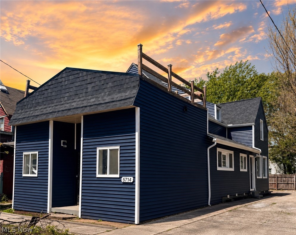 Dark blue house with white trim, rooftop deck railing, and paved driveway at sunset.