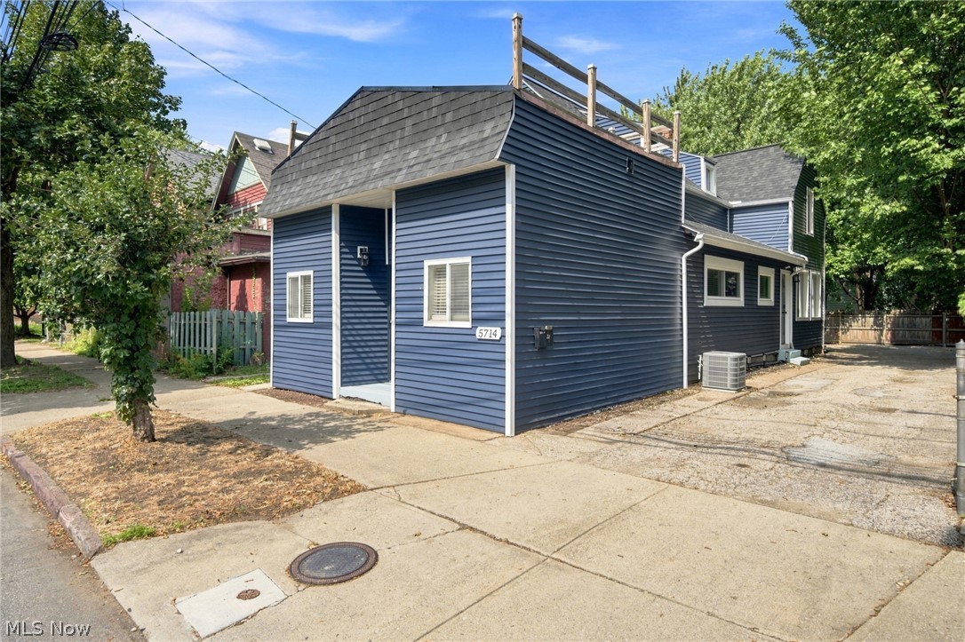 Dark blue-sided house with a raised rooftop deck beside a concrete driveway and mature trees.
