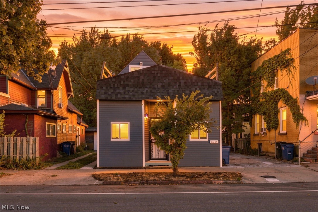 Blue-gray house with a broad shingled roofline at sunset, partly screened by a street-side tree.