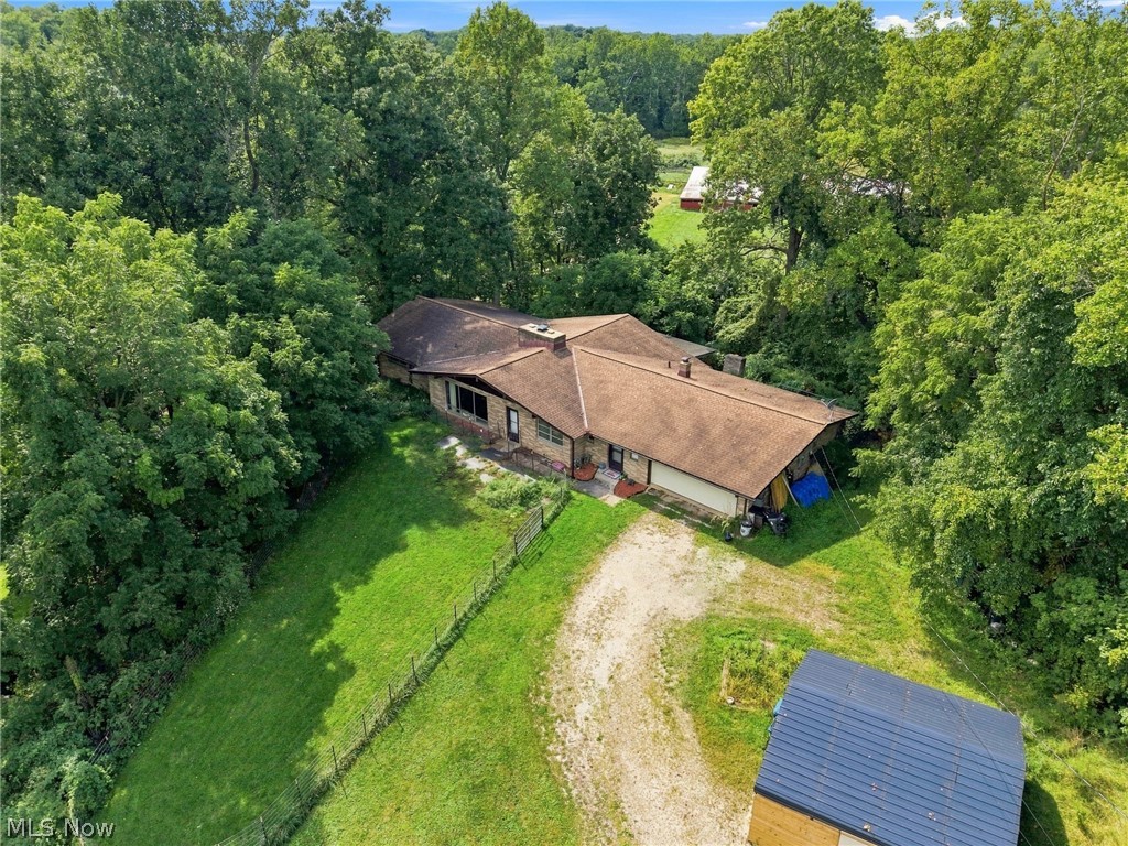 Aerial view of a single-story home surrounded by trees, with a gravel driveway and detached outbuilding.