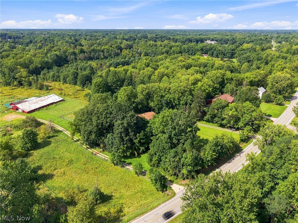Aerial view of wooded acreage with open fields and a long red outbuilding beside a road.