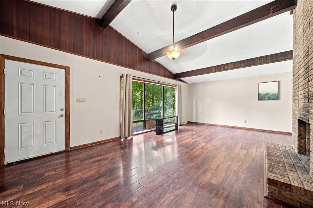Empty living room with exposed ceiling beams, dark wood floors, sliding glass doors, and a brick fireplace.
