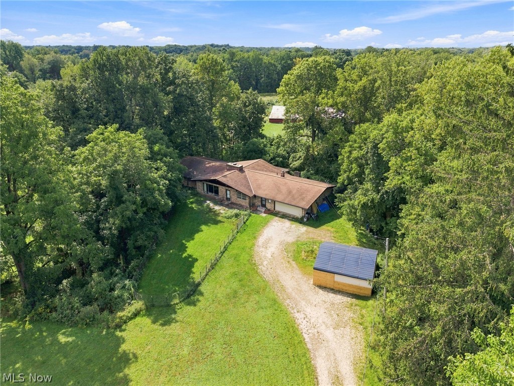 Aerial view of a single-story home, gravel driveway, and solar-panel outbuilding among dense trees.