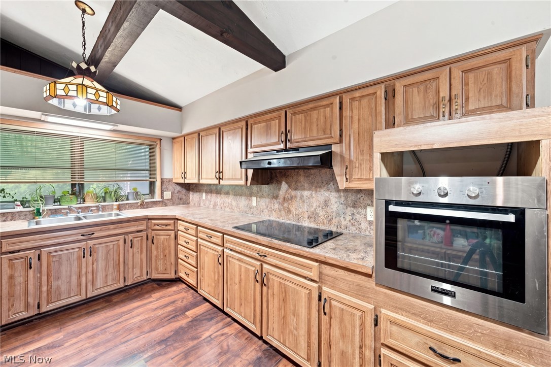 Kitchen with honey-toned wood cabinets, stone-pattern counters, stainless steel oven, and exposed ceiling beams.